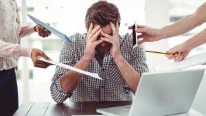 Stressed office worker with hands covering face surrounded by multiple colleagues pointing documents and devices showing stress at work compensation UK 2025 workplace pressure and hostile environment