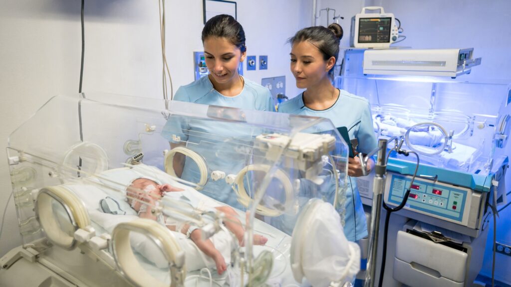 Neonatal intensive care unit showing two healthcare professionals monitoring newborn in incubator with specialized medical equipment for birth injury compensation UK 2025 medical negligence claims