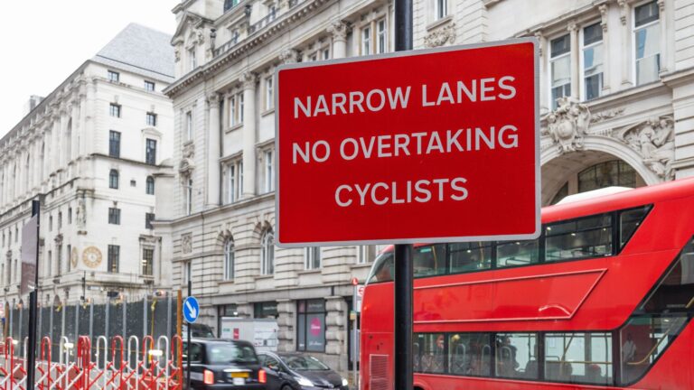 London Road Safety Warning Sign Stating Narrow Lanes No Overtaking Cyclists With Red Bus Representing Cycling Accident Claims London 2026 Borough Liability And Highway Authority Legal Duties